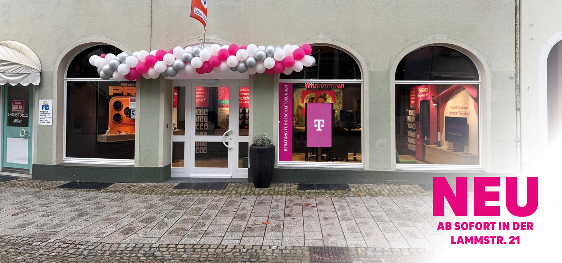 Eine Schaufensterfront mit rosafarbenen, weißen und grauen Luftballons über dem Eingang, die das Branding der Telekom zeigen. Große Fenster geben den Blick auf elektronische Produkte im Inneren frei. Der Text auf dem Bild lautet: "NEU AB SOFORT IN DER LAMMSTR. 21.