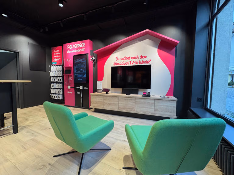 Ein moderner Tele-Planet-Ausstellungsraum mit zwei grünen Stühlen vor einem an der Wand montierten Fernseher in einem rosa-weißen hausförmigen Display mit Produktregalen, einem digitalen Bildschirm und Holzfußboden.