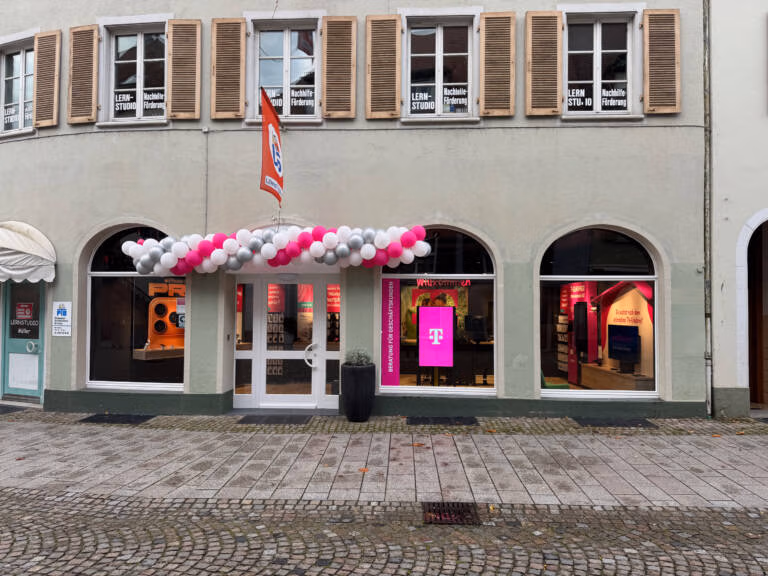 Eine Schaufensterfront mit gewölbten Fenstern ist über dem Eingang mit rosa und weißen Luftballons geschmückt. Innen ist ein helles magentafarbenes Tele-Planet-Schild zu sehen. Die kopfsteingepflasterte Straße draußen führt an Fenstern vorbei, über denen "Lern Studio" steht.