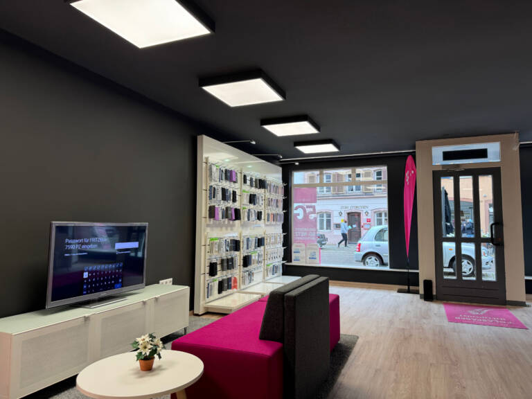 Modernes Tele-Planet-Mobilfunkgeschäft mit schwarzen Wänden, einer Auslage mit Telefonzubehör, einem Fernseher an der linken Wand, rosa Sitzgelegenheiten und großen Glasfenstern mit Blick auf die Straße. Eine kleine Pflanze steht auf einem runden Tisch.