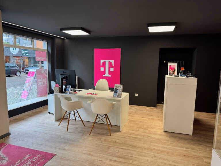 Ein modernes Tele-Planet-Einzelhandelsgeschäft mit einem weißen Schreibtisch, zwei Stühlen, einem Computermonitor und einem großen magentafarbenen T-Mobile-Logo an der Rückwand. Große Fenster geben den Blick auf eine Straße mit Geschäften frei.