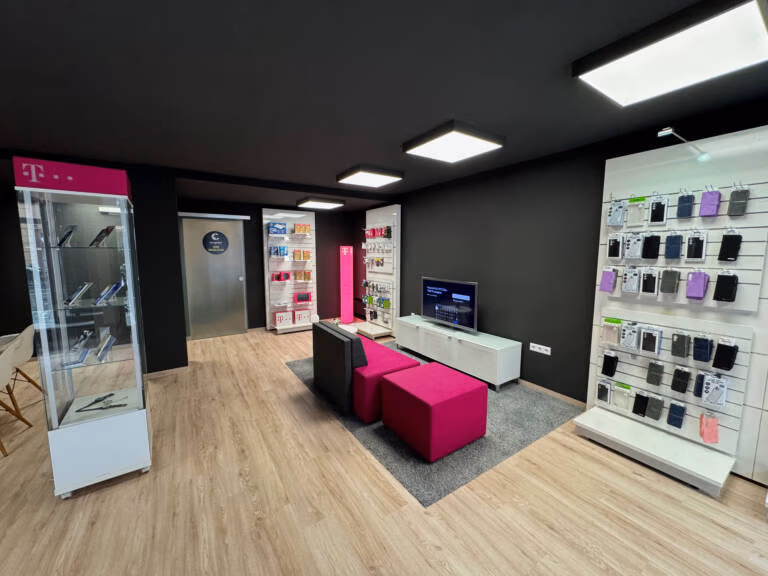 Tele-Planet ist ein modernes Geschäft für Mobiltelefone mit Displays für Telefone und Zubehör, einem Fernseher an der Wand und magentafarbenen und schwarzen Sitzgelegenheiten auf einem grauen Teppich. Die Einrichtung besteht aus schwarzen Wänden und hellen Holzböden.