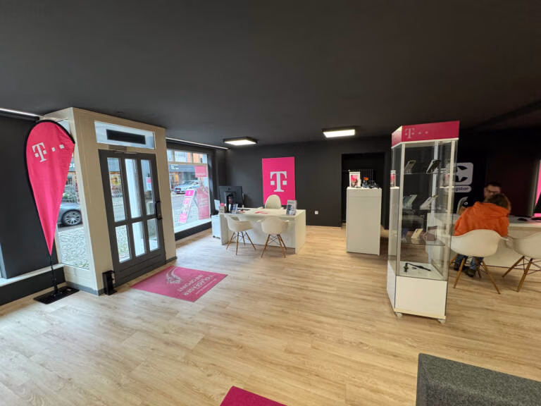 Ein modernes Tele-Planet-Telefongeschäft mit Holzböden, schwarzen Wänden und rosa T-Mobile-Branding. Glastüren lassen natürliches Licht herein. Für den Kundenservice sind Schreibtische und Stühle aufgestellt, und im hinteren Bereich sitzen zwei Personen.