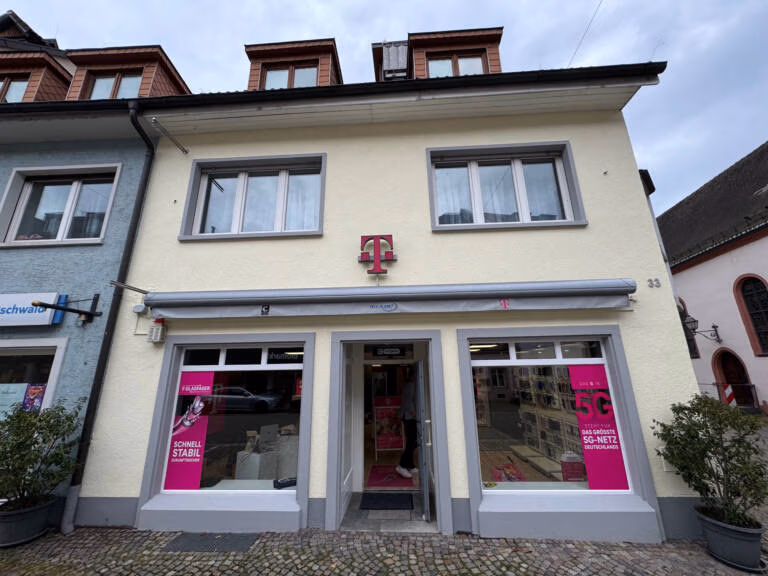 Ein Telekom-Geschäft mit der Marke Tele-Planet hat eine cremefarbene Fassade mit grauer Umrandung und dem magentafarbenen "T"-Logo über dem Eingang. In den großen Fenstern sind Werbeplakate zu sehen. Das Gebäude hat zwei Obergeschosse mit Dachgauben.