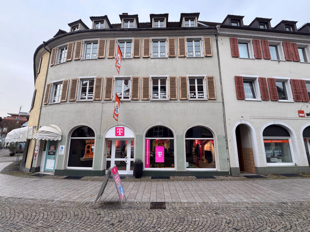 Straßenansicht eines geschwungenen, dreistöckigen Gebäudes mit beigen und weißen Wänden, braunen Fensterläden und einem Tele-Planet Telekom-Geschäft im Erdgeschoss. Der kopfsteingepflasterte Bürgersteig ist in der Nähe des Eingangs mit Schildern versehen, und die Schaufenster tragen rosa Telekom-Logos.