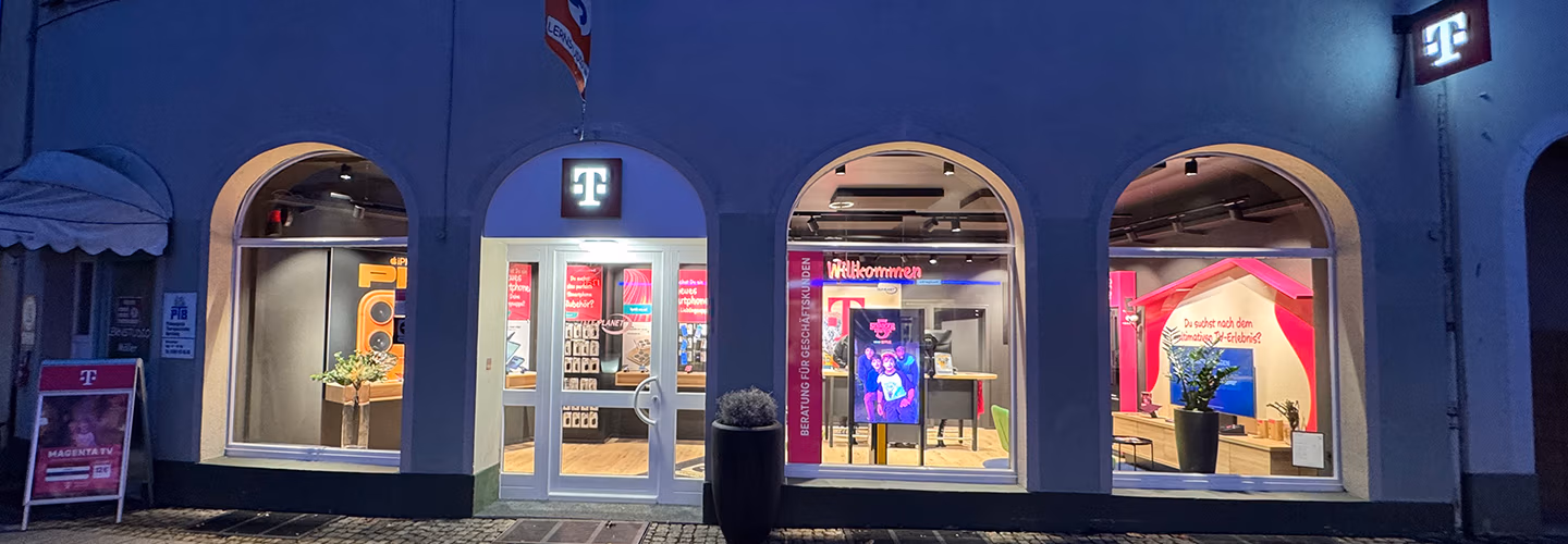 Ein Telekom-Laden mit drei Rundbogenfenstern und einer Glastür, innen hell beleuchtet mit Displays, einem Serviceschalter und digitalen Bildschirmen. Das magentafarbene Telekom-Logo ist über dem Eingang und auf Schildern zu sehen.