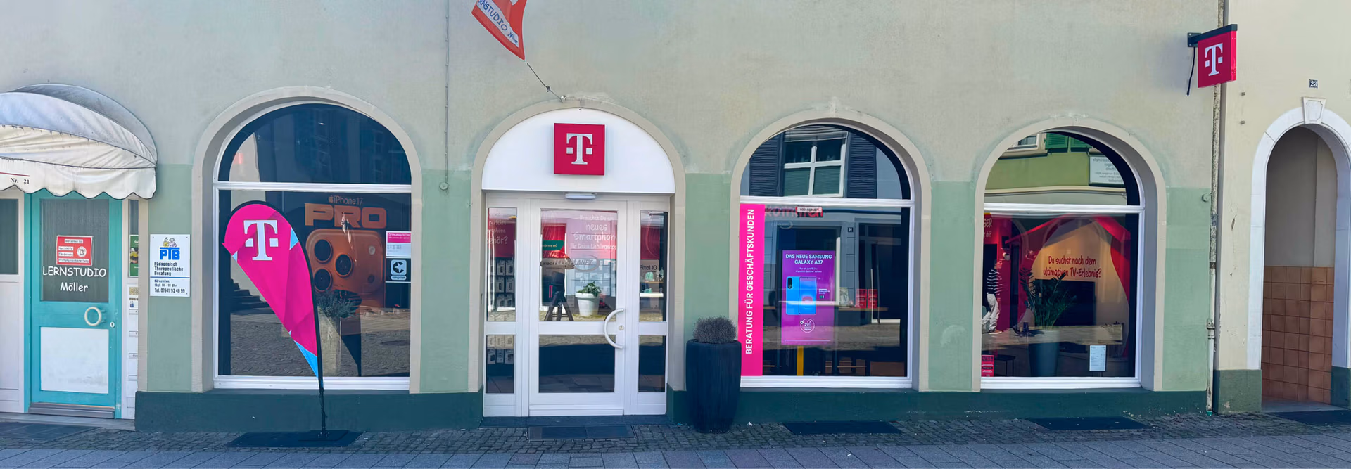 Schaufenster eines Telekom-Shops mit drei gro&szlig;en Bogenfenstern, einer Glast&uuml;r, rosa Telekom-Logos &uuml;ber der T&uuml;r und den Fenstern sowie Werbeplakaten und -bannern innerhalb und au&szlig;erhalb des Gesch&auml;fts.