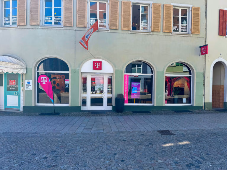 Ein Telekom-Laden mit großen Bogenfenstern und rosafarbener Beschriftung auf einem blassgrünen Gebäude. Der Eingang zeigt das Telekom-Logo über der Tür und Fahnen, und durch die Fenster ist Werbung zu sehen.